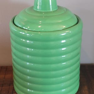 Puede incluir: Un tarro cilíndrico de cerámica de color verde claro con un acabado brillante y una tapa a juego. El cuerpo del tarro presenta una serie de nervaduras horizontales en relieve que rodean todo el contorno, rematado con un asa tipo pomo.
