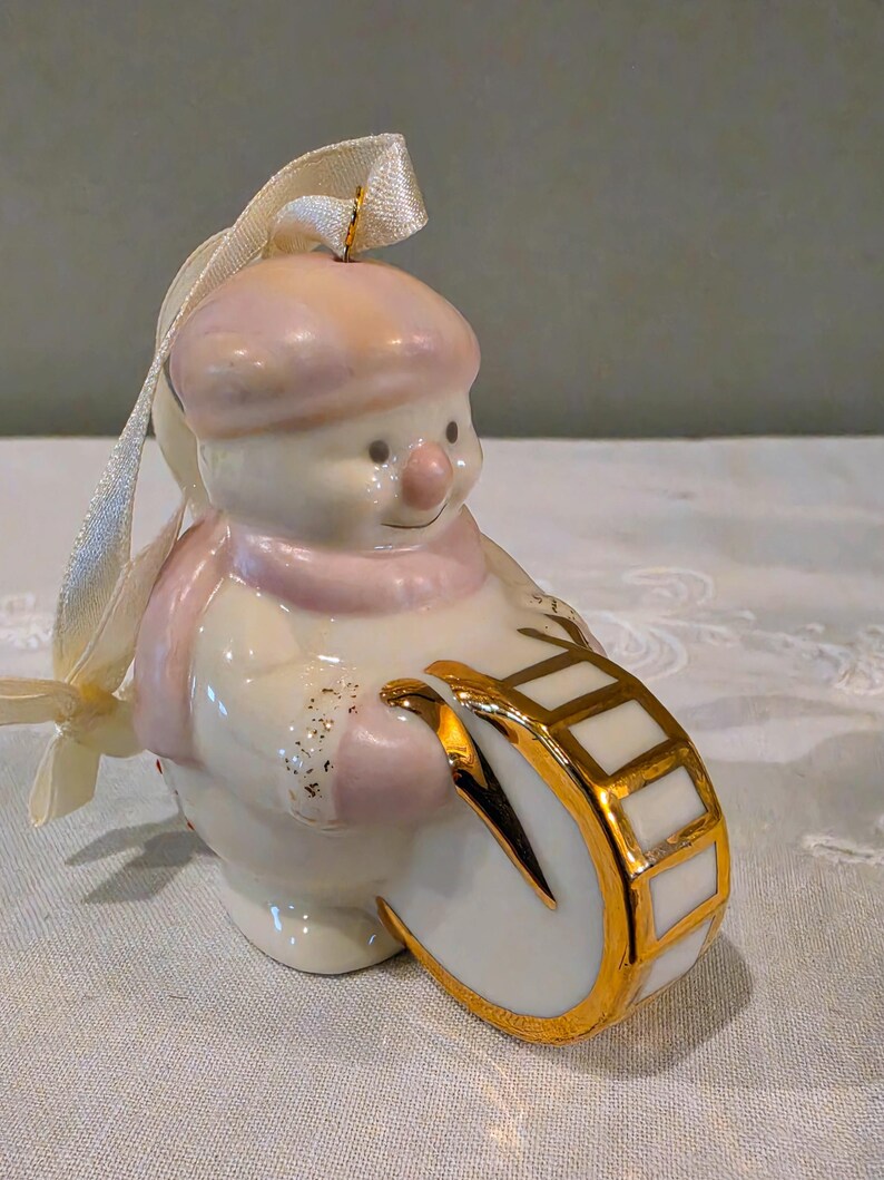 May include: A porcelain snowman ornament wearing a pale pink hat and scarf, playing a white drum with gold trim. The figure features a glossy finish, a small pink nose, and a cream-coloured ribbon loop attached to the top for hanging purposes.