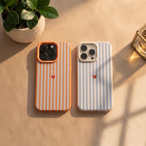 Könnte beinhalten: Zwei Smartphone-Hüllen mit vertikalen Streifen und einem kleinen roten Herz in der Mitte auf der Rückseite. Die linke Hülle hat orange-weiße Streifen mit orangefarbenem Kamerarahmen, die rechte hellblau-weiße Streifen mit cremefarbenem Rahmen.