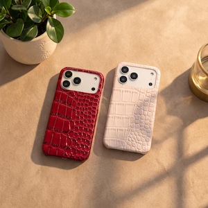 Op de afbeelding: Twee smartphonehoesjes met een gestructureerd krokodillenleerpatroon. Het ene hoesje is dieprood en glanzend, het andere is zacht, mat lichtroze. Beide hoesjes hebben een rechthoekige uitsparing aan de bovenkant voor het camerasysteem.