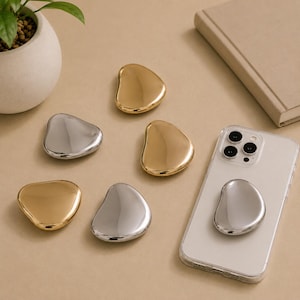 May include: A collection of five smooth, pebble-shaped phone grips in polished gold and silver finishes. Four grips are arranged on a flat surface, while one silver grip is attached to the back of a clear smartphone case with a camera array.