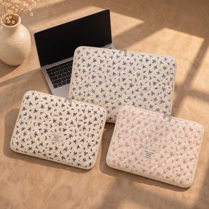 May include: Three rectangular quilted laptop sleeves featuring floral patterns in blue, black, and pink on cream backgrounds. Each sleeve has a zipper closure and a small rectangular label with the text make your day, together, by leece.