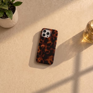Peut inclure: Une coque de smartphone rectangulaire arborant un motif écaille de tortue marron et noir. La coque présente une découpe pour un système de triple objectif photo dans le coin supérieur gauche, révélant les objectifs et le flash de l'appareil.