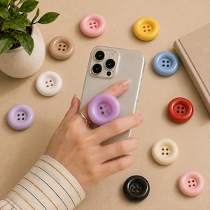 Può includere: Uno smartphone con un supporto a forma di bottone viola attaccato sul retro, circondato da vari bottoni sparsi nei colori rosa, giallo, blu, rosso, nero, bianco, beige e lavanda, disposti su una superficie piana.