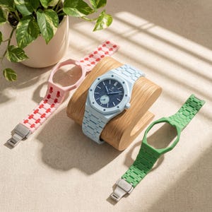 May include: A light blue wristwatch with a textured dark blue dial and a small sub-dial, mounted on a wooden stand. Two additional watch bands are present: one pink with red accents and one solid green, both featuring metal clasps.