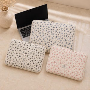 Könnte beinhalten: Drei rechteckige, gesteppte Laptoptaschen mit Blumenmustern in Blau, Schwarz und Rosa auf cremefarbenem Hintergrund. Jede Tasche hat einen Reißverschluss und ein kleines Stoffetikett mit dem Text "make your day happier by kazun".