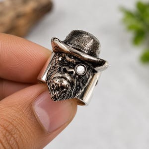 May include: A silver-toned metal ring featuring a detailed sculpted gorilla head wearing a textured fedora hat. The gorilla has a single clear round stone set into one eye, resembling a monocle, with dark oxidized detailing to highlight the features.
