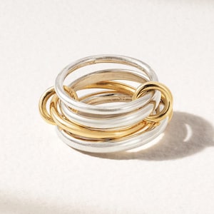 Puede incluir: Un anillo de varias bandas que presenta una combinación de bandas de metal pulido en tonos plateados y dorados. El diseño consta de varios anillos circulares entrelazados de diferentes grosores que están conectados para formar una sola pieza.