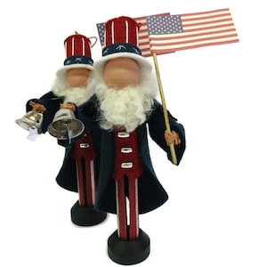 May include: Two patriotic wooden figures dressed in red, white, and blue with a white beard and a top hat. One figure holds two silver bells and the other holds a small American flag.