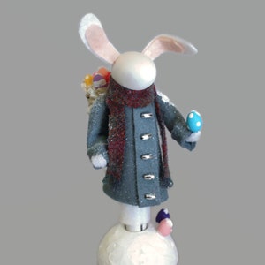 Peut inclure: Un lapin blanc portant un manteau bleu avec une écharpe rouge et tenant un œuf bleu. Le lapin est debout sur une base blanche avec quelques œufs colorés autour de lui.