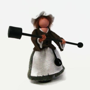 May include: A handmade doll wearing a brown dress, white apron, and white bonnet. The doll is holding a black stick with a black ball on the end.