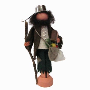 May include: A wooden figurine of a person wearing a brown coat, green pants, and a silver hat. The figurine is holding a stick and a bag with fruit and a small box. The figurine is standing on a wooden base.