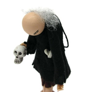 May include: A handmade doll with a white wig, black dress, and a skull in its hand. The doll has a stitched-up face and a bone in its pocket.