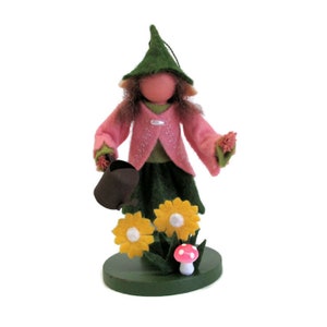 May include: A felt gnome doll wearing a pink jacket, green skirt, and green hat, holding a watering can and standing in front of yellow flowers and a red mushroom.