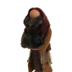 May include: A handcrafted figurine of a person, likely a shepherd, with a wooden head and a felted wool outfit. The figure wears a burgundy shawl, a brown coat, and holds a black bundle. The base is a terracotta color.