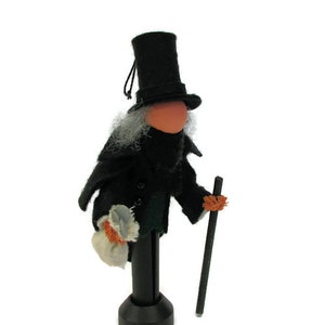 May include: A black felt doll wearing a top hat, a black coat, and holding a walking stick. The doll has a white beard and is holding a small white bag.