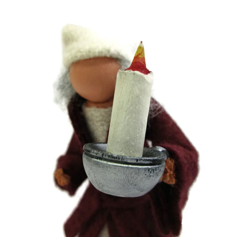 Pu&ograve; includere: Una piccola bambola con un cappello bianco e un vestito color borgogna tiene una candela in un portacandele argentato. La candela &egrave; bianca con una fiamma rossa e gialla.