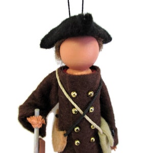 May include: A felt doll wearing a brown coat with gold buttons, a black hat, and a white rope belt. The doll is holding a wooden stick.