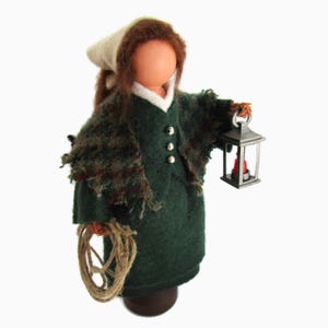 May include: A handcrafted doll dressed in a dark green felt dress and shawl, holding a small metal lantern with a red candle. The doll has brown hair, a white cap, and a rope in the other hand. The doll is standing on a dark brown base.