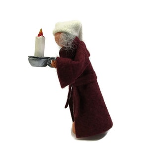 Pu&ograve; includere: Una bambola di feltro che indossa una veste color borgogna e un berretto da notte bianco, tiene un piccolo vassoio di metallo con una candela accesa.