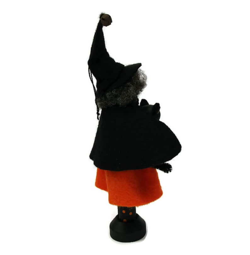 May include: A black witch doll with a black hat and a black cat in her arms. She is wearing an orange skirt with black polka dots and is standing on a black base.