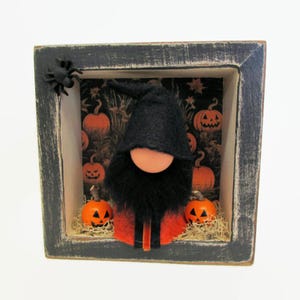 May include: A Halloween shadow box with a gnome wearing a black hat and beard, an orange nose, and an orange and black outfit. The background features a pumpkin pattern. Two carved pumpkins are in front of the gnome, and a black spider is on the frame.