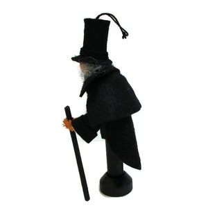May include: A black felt doll wearing a top hat and a long black coat, holding a stick. The doll is standing on a wooden base.