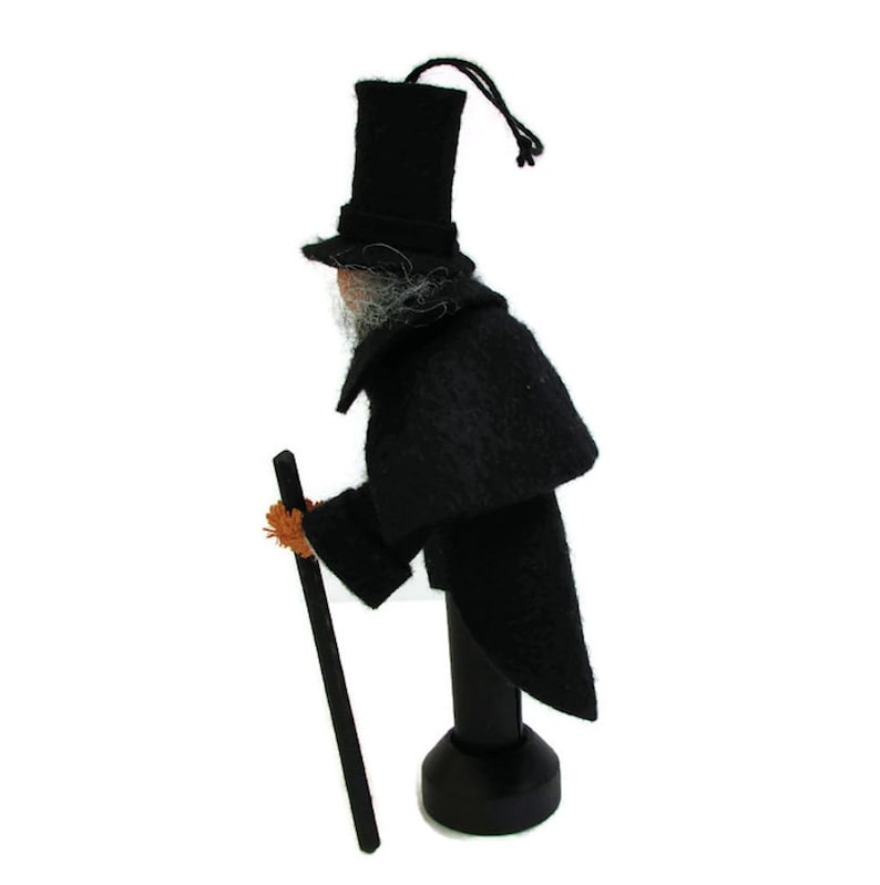 May include: A black felt doll wearing a top hat and a long black coat, holding a stick. The doll is standing on a wooden base.