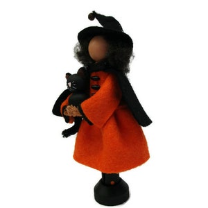 May include: A black witch doll with a black hat and orange dress holds a black cat. The doll is standing on a black base.