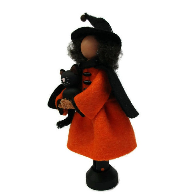 May include: A black witch doll with a black hat and orange dress holds a black cat. The doll is standing on a black base.