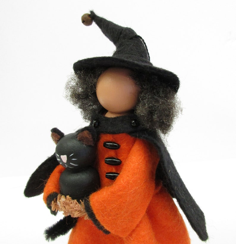 May include: A witch doll with black hair and a black hat holds a black cat doll. The witch doll is wearing an orange dress with black buttons and a black cape.