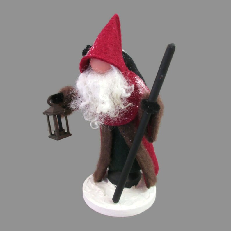 May include: A handmade felt gnome figurine with a red hat, brown fur coat, and white beard. The gnome is holding a lantern and a wooden staff. The gnome is standing on a white base that looks like snow.