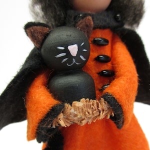 May include: A felt witch doll wearing a black cape and orange dress, holding a black cat doll with painted white facial features.