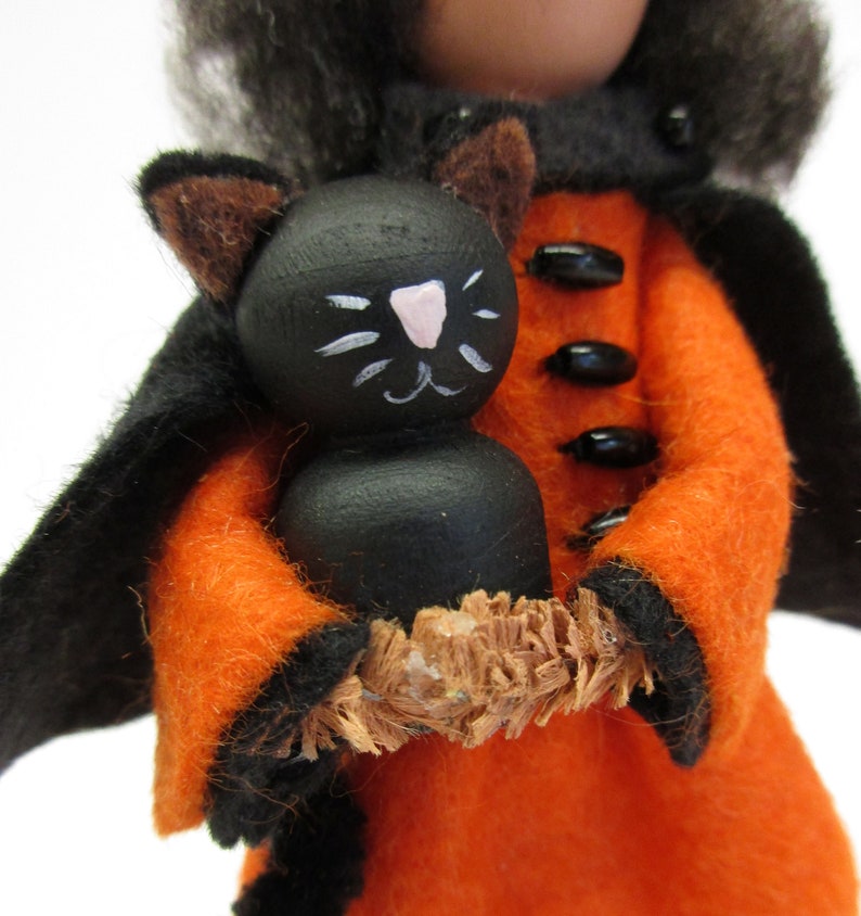 May include: A felt witch doll wearing a black cape and orange dress, holding a black cat doll with painted white facial features.