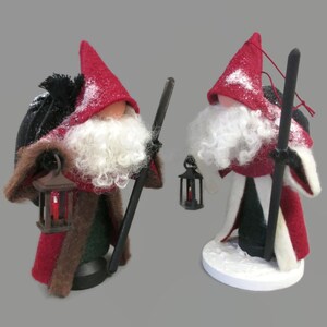 May include: Two felt Christmas gnomes with red hats, white beards, and black staffs. Each gnome holds a small lantern with a candle inside. The gnomes are standing on a white base.