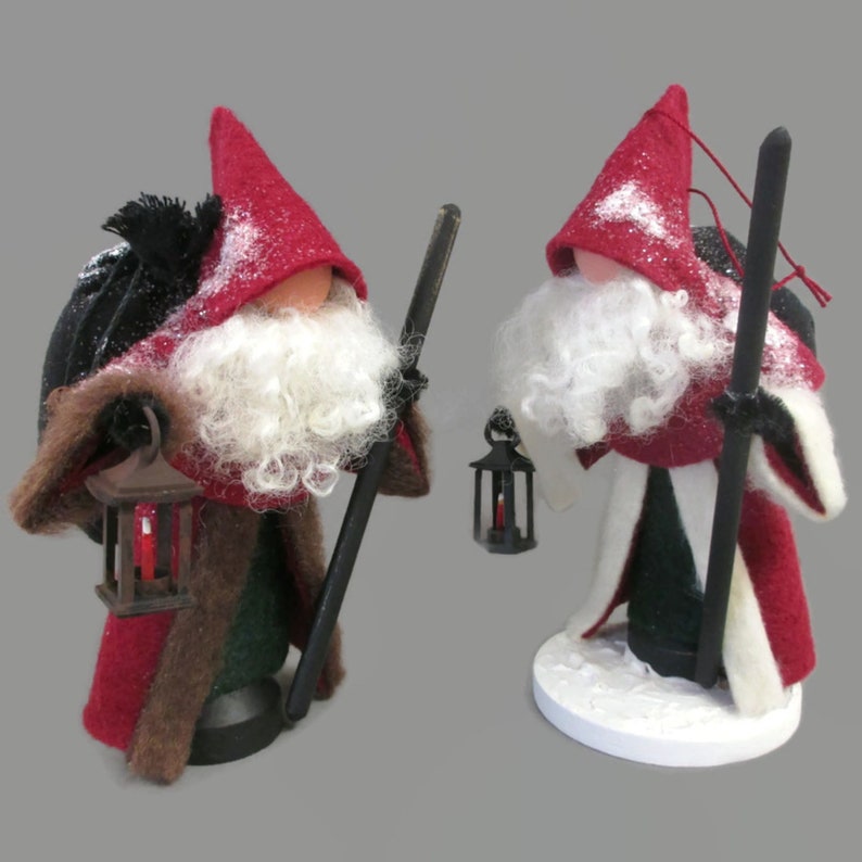 May include: Two felt Christmas gnomes with red hats, white beards, and black staffs. Each gnome holds a small lantern with a candle inside. The gnomes are standing on a white base.