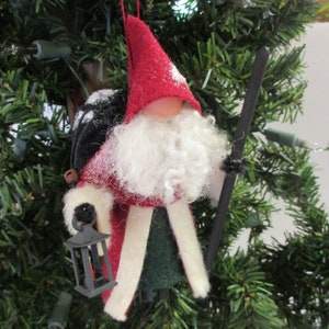 May include: A felt Christmas ornament depicting a Santa Claus figure with a white beard, red hat, and green and white robe. He is carrying a lantern and a stick.