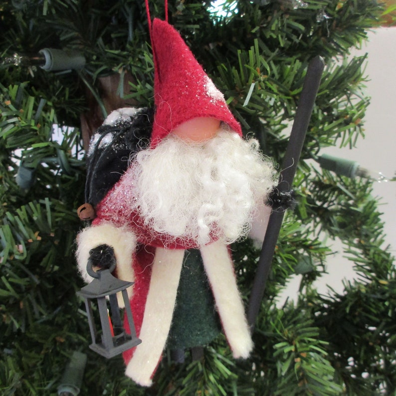 May include: A felt Christmas ornament depicting a Santa Claus figure with a white beard, red hat, and green and white robe. He is carrying a lantern and a stick.