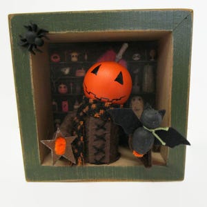 May include: A Halloween-themed shadow box with a carved orange pumpkin head, a black and orange scarf, and a brown body. A black bat with green accents and a star are also featured. The box has a green frame and a spider.