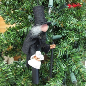 May include: A felt Christmas ornament depicting a man wearing a black top hat, a black coat, and a white beard. He is holding a walking stick and a small white package. The ornament is hanging on a green artificial Christmas tree.