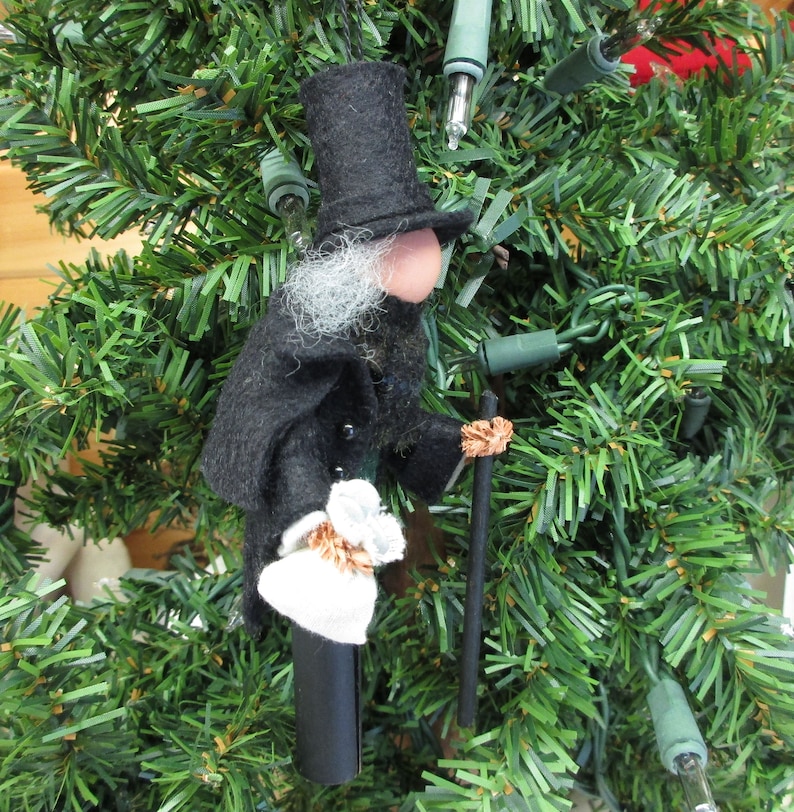 May include: A felt Christmas ornament depicting a man wearing a black top hat, a black coat, and a white beard. He is holding a walking stick and a small white package. The ornament is hanging on a green artificial Christmas tree.