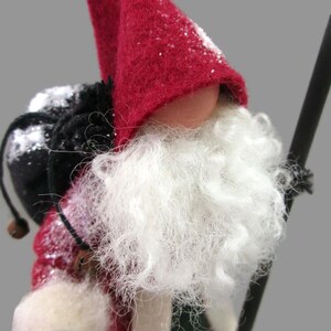 May include: A red felt gnome with a white beard and a black bag on its back. The gnome is wearing a red hat with white snow on it.