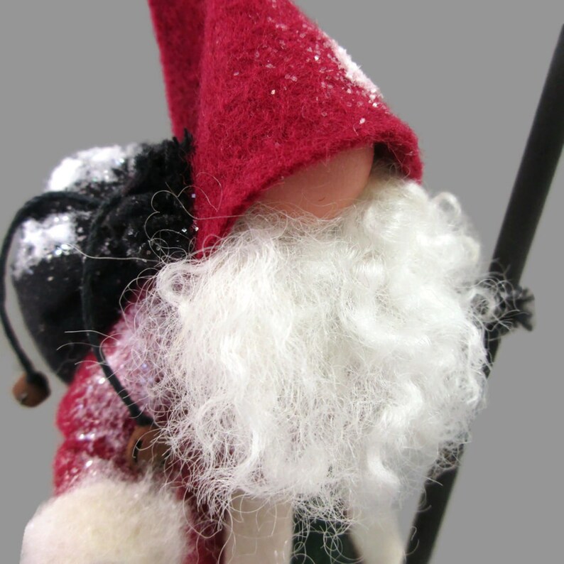 May include: A red felt gnome with a white beard and a black bag on its back. The gnome is wearing a red hat with white snow on it.