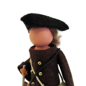 May include: A brown felt doll wearing a black felt beret and a brown felt coat with gold buttons. The doll has a wooden head and a painted face.