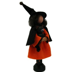 May include: A black witch doll with a black hat and orange dress holds a black cat. The doll is standing on a black and white polka dot base.