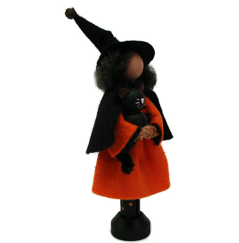 May include: A black witch doll with a black hat and orange dress holds a black cat. The doll is standing on a black and white polka dot base.