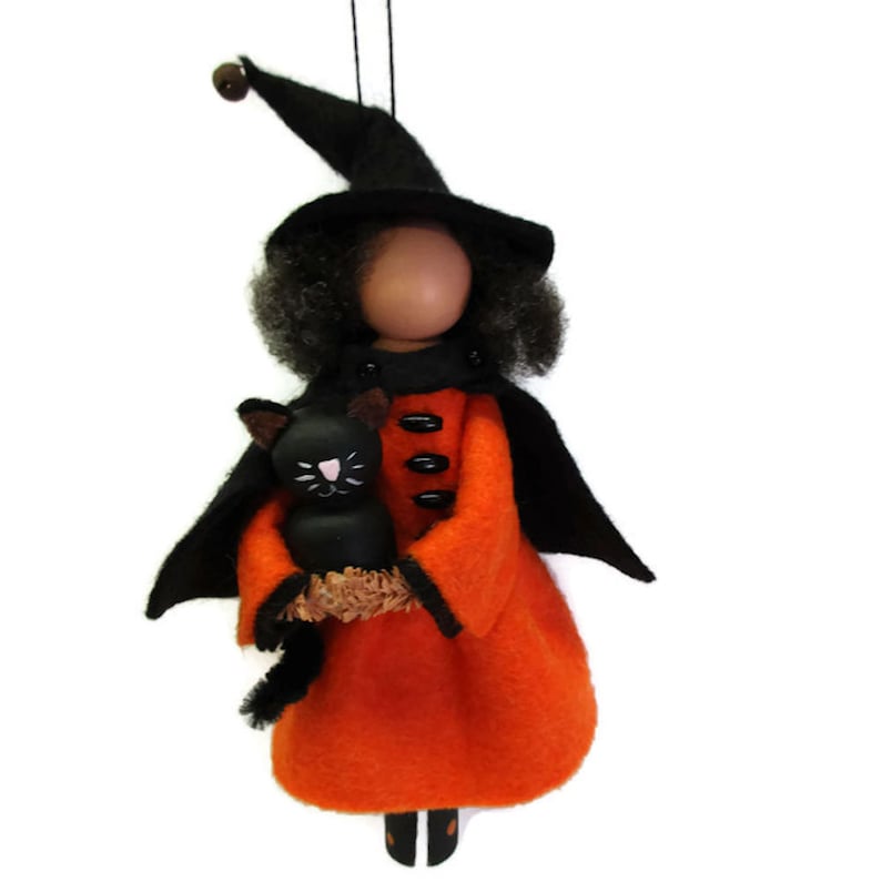 May include: A handmade witch doll ornament with black hair, a black hat, and an orange dress. She is holding a black cat in her arms.
