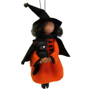 May include: A handmade Halloween ornament featuring a witch doll with black hair, a black hat, and an orange dress. She is holding a black cat in her arms.