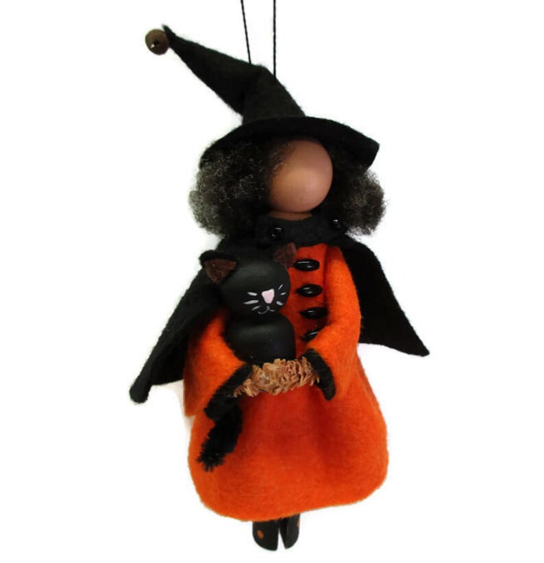May include: A handmade Halloween ornament featuring a witch doll with black hair, a black hat, and an orange dress. She is holding a black cat in her arms.
