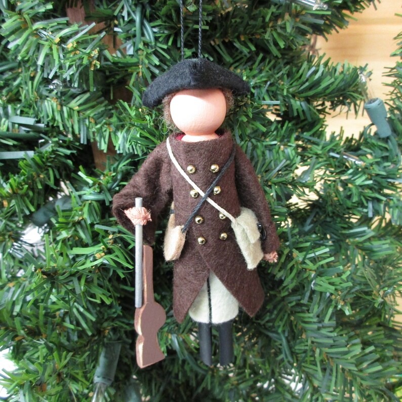 May include: A felt doll ornament of a soldier wearing a brown coat, black hat, and holding a wooden rifle. The doll is decorated with gold buttons and white accents.